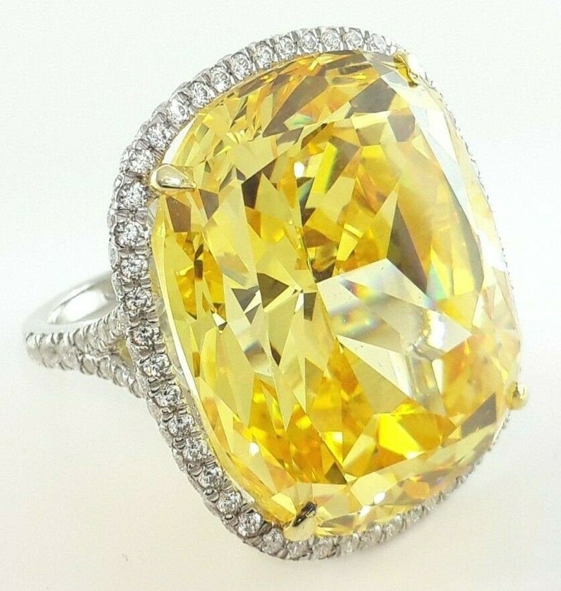 May include: A large, eye-catching ring featuring a vibrant yellow gemstone. The gemstone is rectangular with a faceted cut, surrounded by a halo of smaller, clear stones. The band is silver-toned, adding to the ring's elegant design.
