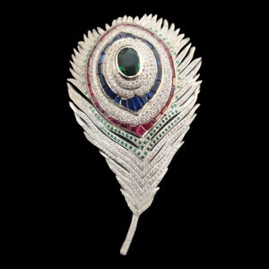 Pode incluir: Um broche de prata em forma de pena de pavão com uma grande esmeralda verde no centro, cercada por safiras azuis e rubis vermelhos menores.
