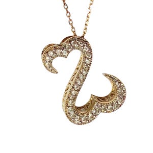 Peut inclure: Un collier pendentif doré avec un motif de cœur ouvert stylisé. Le pendentif est incrusté de petits pierres précieuses scintillantes et est suspendu à une chaîne délicate. Le collier est sur fond blanc.