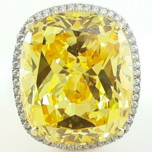 May include: A large, cushion-cut yellow gemstone ring. The vibrant yellow stone is surrounded by a halo of smaller, clear gemstones. The ring's setting appears to be gold or a gold-toned metal.