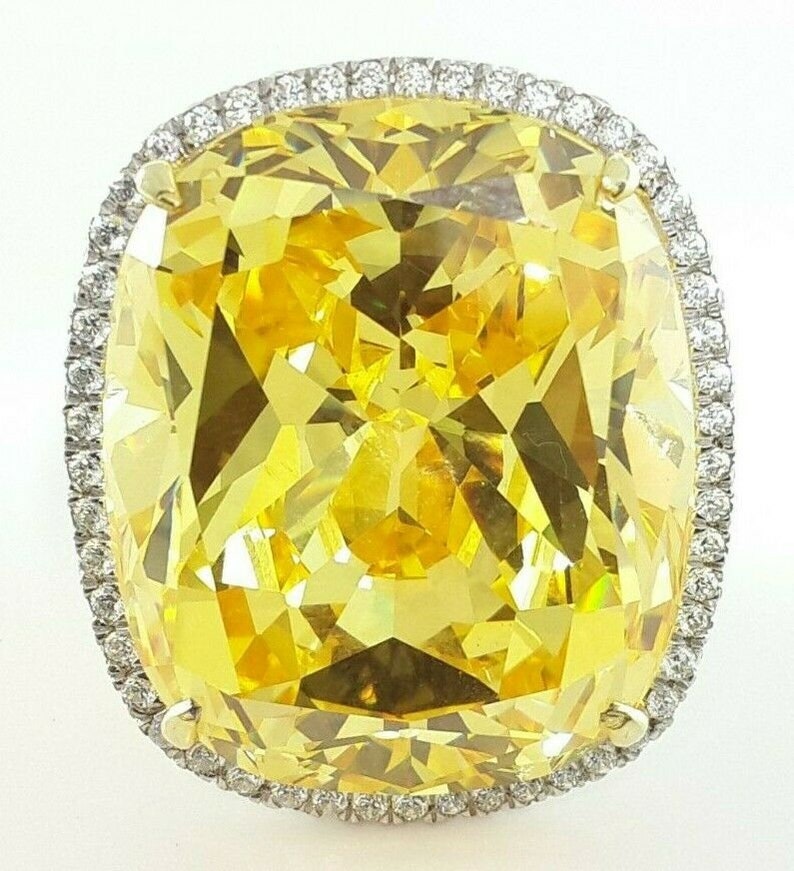 May include: A large, cushion-cut yellow gemstone ring. The vibrant yellow stone is surrounded by a halo of smaller, clear gemstones. The ring's setting appears to be gold or a gold-toned metal.