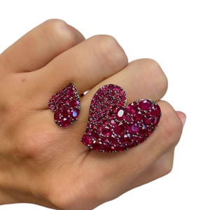 May include: Two heart-shaped rings, one large and one small, are adorned with numerous small, deep red gemstones. The rings are set on a hand against a white background. The larger heart ring is on the index finger, and the smaller ring is on the ring finger.