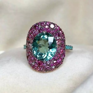 Può includere: Un anello con una grande pietra preziosa verde di forma ovale circondata da un doppio alone di pietre preziose rosa e bianche.