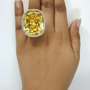 May include: A large, square-cut yellow gemstone ring with a halo of small, clear stones. The ring is set on a hand with a neutral skin tone, against a white background. The ring's setting appears to be gold or a gold-toned metal.