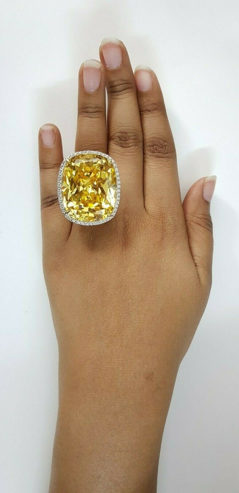 May include: A large, square-cut yellow gemstone ring with a halo of small, clear stones. The ring is set on a hand with a neutral skin tone, against a white background. The ring's setting appears to be gold or a gold-toned metal.