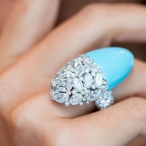 May include: A ring with a large, oval-shaped turquoise stone set in a bed of sparkling diamonds. The ring is designed to fit over the fingernail.