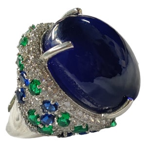 Puede incluir: Un anillo de plata con una gran piedra ovalada azul cabujón rodeada de piedras más pequeñas de color verde y azul.
