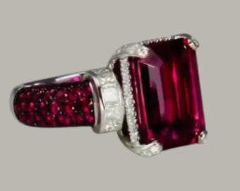 May include: A silver ring featuring a large, rectangular, deep red gemstone as the focal point. The ring's band is adorned with smaller, round red gemstones, and the setting is embellished with clear, sparkling accent stones.