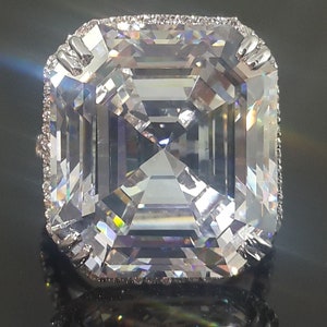 May include: A large, clear, square-shaped diamond ring with a silver band and a row of small diamonds around the edge of the setting.