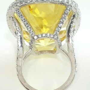 May include: A large, yellow gemstone ring with a square-cut center stone. The ring features a halo of small, clear stones around the center and along the band. The setting is silver-toned metal.
