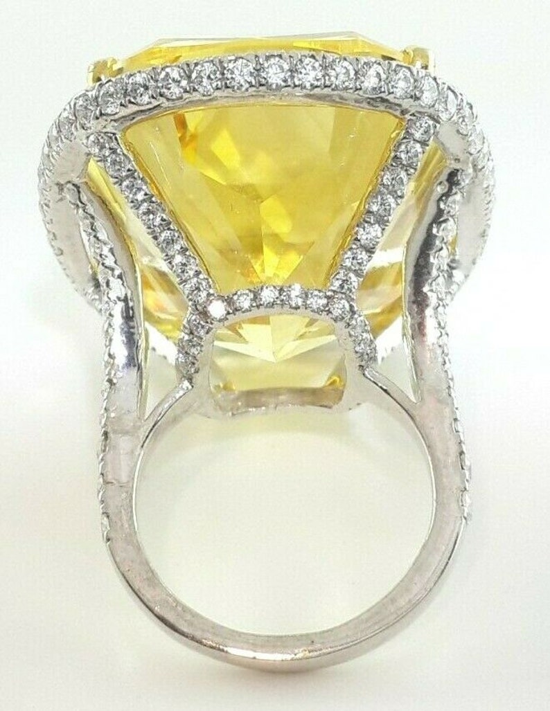 May include: A large, yellow gemstone ring with a square-cut center stone. The ring features a halo of small, clear stones around the center and along the band. The setting is silver-toned metal.