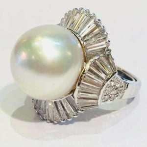 May include: A white pearl ring featuring a large, round pearl set in a white gold band. The band is adorned with a cluster of baguette-cut diamonds.