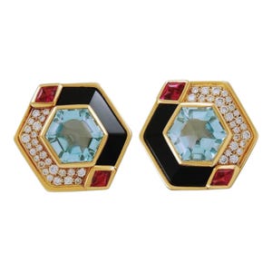 May include: A pair of gold earrings with a hexagonal design. Each earring features a blue gemstone surrounded by a black enamel border and a ring of small diamonds. The earrings are accented with red gemstones.
