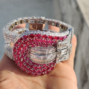 May include: A silver bracelet with a large, red ruby-encrusted buckle. The bracelet is set with smaller diamonds around the buckle and the band.