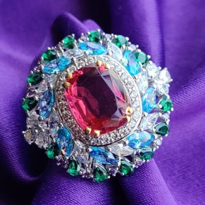 May include: A large, ornate ring featuring a central oval-cut, deep pink gemstone. The ring is surrounded by a halo of smaller, multi-colored gemstones in shades of blue, green, and white, set in a silver-toned metal. The ring is displayed on a purple fabric.