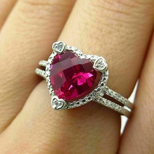 May include: A heart-shaped ring with a large, bright pink gemstone surrounded by a halo of smaller, clear gemstones. The ring is set in white metal and has a delicate band.