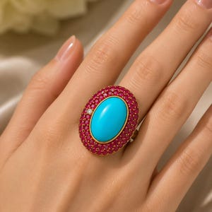 Peut inclure: Une bague en or avec une grande pierre centrale turquoise ovale et lisse. La pierre est entourée d'une large bordure composée de nombreuses petites rubis ronds rose foncé sertis dans l'or, avec un petit diamant rond et clair en accent.