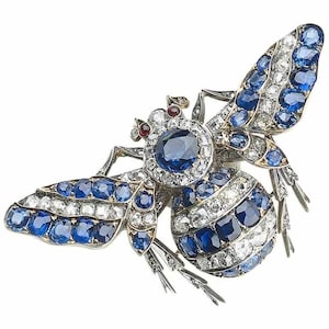 Può includere: Spilla a forma di ape con dettagli intricati. La spilla presenta una grande gemma blu centrale circondata da gemme bianche più piccole. Le ali e il corpo sono adornati con gemme blu e bianche.
