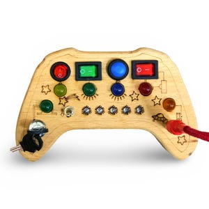 May include: A wooden toy game controller with a variety of buttons, switches, and lights. The controller is shaped like a traditional video game controller and has a red, blue, and green color scheme.