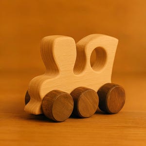 May include: A wooden toy train with a light brown body and four dark brown wheels. The train has a simple design with two rounded openings in the front.