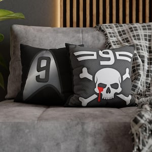 May include: Two black throw pillows with unique designs. One features a silver number 9 on a black background. The other has a skull and crossbones design with the number 95, and a red tear. The pillows are on a gray couch.
