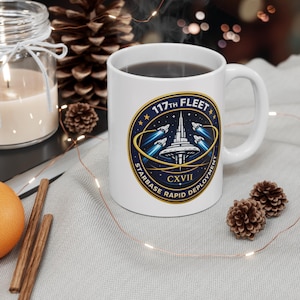 May include: White ceramic mug with a black beverage inside. The mug features a circular emblem with the text "117th Fleet Starbase Rapid Deployment CXVII" and a graphic of a space station and rockets. The mug is on a light gray surface.