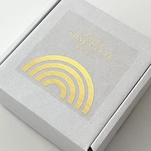 May include: A white box with a clear sticker featuring a gold rainbow design. The sticker has the words "SUNCATCHER" and "Rainbow" and "MARU HOME" printed in gold. The rainbow design is made up of several curved lines.