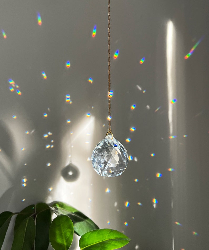 May include: A clear, faceted crystal ball hanging from a gold chain, casting rainbow light patterns on a gray wall. The crystal is teardrop-shaped, reflecting sunlight. Green leaves are visible in the lower left corner.