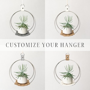 May include: Four hanging planters with a white ceramic pot holding a green plant. Each planter has a silver metal ring, a woven base, and a leather or metal top loop. The text "CUSTOMIZE YOUR HANGER" is displayed below the planters.