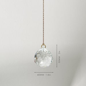 May include: A clear, faceted crystal ball, 1.6 inches wide and 2 inches long, hangs from a gold-colored chain. The crystal sphere is suspended against a plain white background, showcasing its geometric design and reflective qualities.