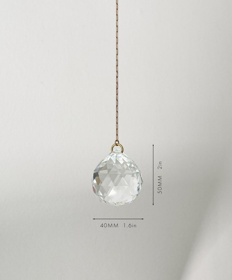 May include: A clear, faceted crystal ball, 1.6 inches wide and 2 inches long, hangs from a gold-colored chain. The crystal sphere is suspended against a plain white background, showcasing its geometric design and reflective qualities.