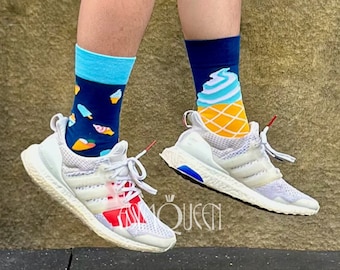 May include: A pair of white sneakers with blue and red accents. The sneakers are worn by a person wearing blue denim shorts and socks with ice cream designs. One sock features a blue ice cream cone with a yellow waffle cone and the other sock features a blue and pink ice cream sandwich.
