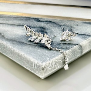 Puede incluir: Un par de pendientes de plata con un diseño de hoja y cristales transparentes. Los pendientes están sobre una superficie de mármol gris.