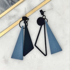 Puede incluir: Un par de pendientes geométricos con un marco de metal negro y triángulos de madera azul. Los pendientes tienen un diseño simple y moderno.