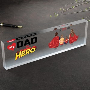 K&ouml;nnte beinhalten: Ein transparenter Acrylblock mit dem Text "My Dad is Hero" in einer Comic-Schriftart. Der Block zeigt auch eine f&uuml;nfk&ouml;pfige Familie, die rote Superhelden-Umh&auml;nge tr&auml;gt, mit ihren Namen darunter.