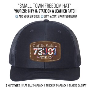 May include: A navy blue trucker hat with a mesh back and a curved brim. The hat features a leather patch with the text "Small Town Freedom" and the zip code 73301, along with "Austin, TX". The hat has the text "3 HAT STYLES I FLAT BILL SNAPBACK I TRUCKER SNAPBACK I CLASSIC DAD HAT".