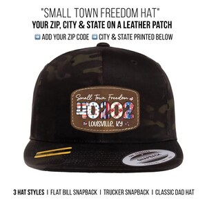 May include: Black and camouflage flat bill snapback hat with a leather patch. The patch displays the text "Small Town Freedom" and the zip code 40102, with "Louisville, KY" below. The hat has a gold accent on the bill.