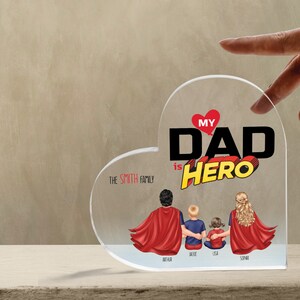 K&ouml;nnte beinhalten: Eine transparente, herzf&ouml;rmige Acrylplatte mit dem Text "My Dad is Hero" in Rot und Schwarz. Der Text ist in einer Comic-Schriftart gehalten. Unter dem Text befinden sich vier Figuren von Personen in roten Umh&auml;ngen, mit dem Text "The Smith Family" dar&uuml;ber. Die Figuren sind mit "Arthur", "Jackie", "Lisa" und "Sophia" beschriftet.
