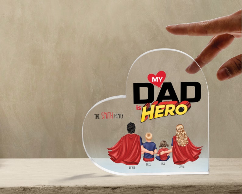K&ouml;nnte beinhalten: Eine transparente, herzf&ouml;rmige Acrylplatte mit dem Text "My Dad is Hero" in Rot und Schwarz. Der Text ist in einer Comic-Schriftart gehalten. Unter dem Text befinden sich vier Figuren von Personen in roten Umh&auml;ngen, mit dem Text "The Smith Family" dar&uuml;ber. Die Figuren sind mit "Arthur", "Jackie", "Lisa" und "Sophia" beschriftet.