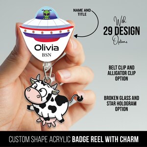 Op de afbeelding: Een op maat gemaakte acryl badge-rolhouder met een bedel. De rolhouder heeft een cartoon UFO met een groene alien erin. De bedel is een cartoon koe met zwarte en witte vlekken. De tekst "Olivia BSN" is op de rolhouder gedrukt.