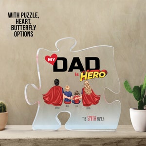 K&ouml;nnte beinhalten: Ein klares Acryl-Puzzleteil in Herzform mit den Worten "My Dad is Hero" in Rot, Schwarz und Wei&szlig;. Das Puzzleteil zeigt eine vierk&ouml;pfige Familie, wobei der Vater einen roten Superhelden-Umhang tr&auml;gt und die Kinder blaue Hemden. Die Namen "Axe", "Jackie", "Lisa" und "Sophia" stehen unter der Familie. Der Text "The Smith Family" steht unter der Familie.