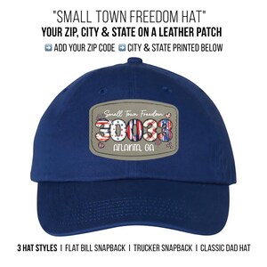 May include: A navy blue baseball cap with a leather patch. The patch displays the text "Small Town Freedom" and the zip code "30038" with "Atlanta, GA" below. The hat has a curved brim.