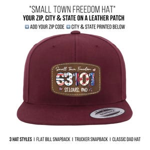 May include: A burgundy flat bill snapback hat with a leather patch. The patch displays the text "Small Town Freedom" and the zip code "63101" with the city "St. Louis, MO". The hat has a classic design.