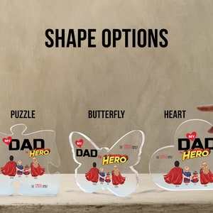 K&ouml;nnte beinhalten: Drei Acrylformen, ein Puzzle, ein Schmetterling und ein Herz, jede mit einem personalisierten Design mit dem Text "Mein Vater ist ein Held" und einer Familienillustration. Die Formen sind klar und haben eine gl&auml;nzende Oberfl&auml;che.
