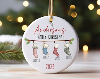May include: White ceramic Christmas ornament with the text "The Andersons Family Christmas" and four stockings hanging from a branch. The stockings are red and white striped, blue and white polka dotted, red and black plaid, and gray and white plaid. The names Gloria, Emily, Jayk, and Louis are written below the stockings. The year 2023 is written at the bottom of the ornament.