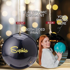 May include: A round, black ceramic ornament with a yellow inscription that reads "Sophia". The back of the ornament features a photo of a woman holding a blue bowling ball.