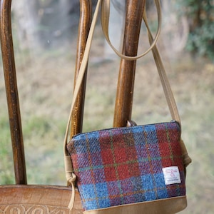 May include: A crossbody bag featuring a red and blue plaid wool fabric with a tan suede base and a matching tan adjustable strap. A small white rectangular label with red text is stitched to the lower right corner of the bag, which has a zipper closure.
