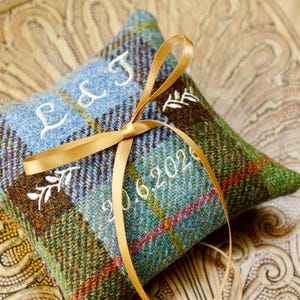 May include: A square ring pillow made of blue, green, and brown plaid fabric. The pillow features white embroidered text reading L & J and 20.6.2020, accented with small embroidered leaf motifs. A thin, gold-colored satin ribbon is tied in a bow.