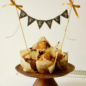 Könnte beinhalten: Ein hölzerner Kuchenständer hält mehrere Muffins in Papierförmchen. Ein Banner mit der Aufschrift "STEVEN" ist zwischen zwei Holzspießen gespannt, die jeweils mit einem goldenen Band verziert sind. Die Muffins scheinen Blaubeeren zu enthalten.