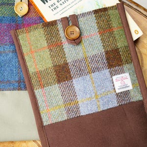 May include: A rectangular fabric sleeve featuring a green, blue, and brown plaid tweed pattern with a solid brown fabric base, a brown button closure, and a white Harris Tweed label. The sleeve is positioned next to a book titled Wonderful Life.